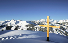 Holzkreuz auf schneebedecktem Gipfel mit beeindruckender Berglandschaft im Hintergrund.