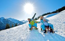 Familie fährt in der Sonne mit Schlitten auf einem verschneiten Hang in Saalbach-Hinterglemm.