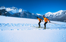 Zwei Skifahrer in orangen Jacken auf verschneiter Piste vor bergiger Kulisse.